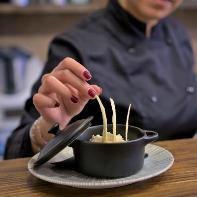 Chef colocando pan crujiente en cazuela negra con crema, fotografía gastronómica profesional en restaurante.
