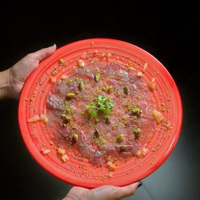Carpaccio gourmet servido en plato rojo con pistachos y brotes verdes, fotografía gastronómica profesional.